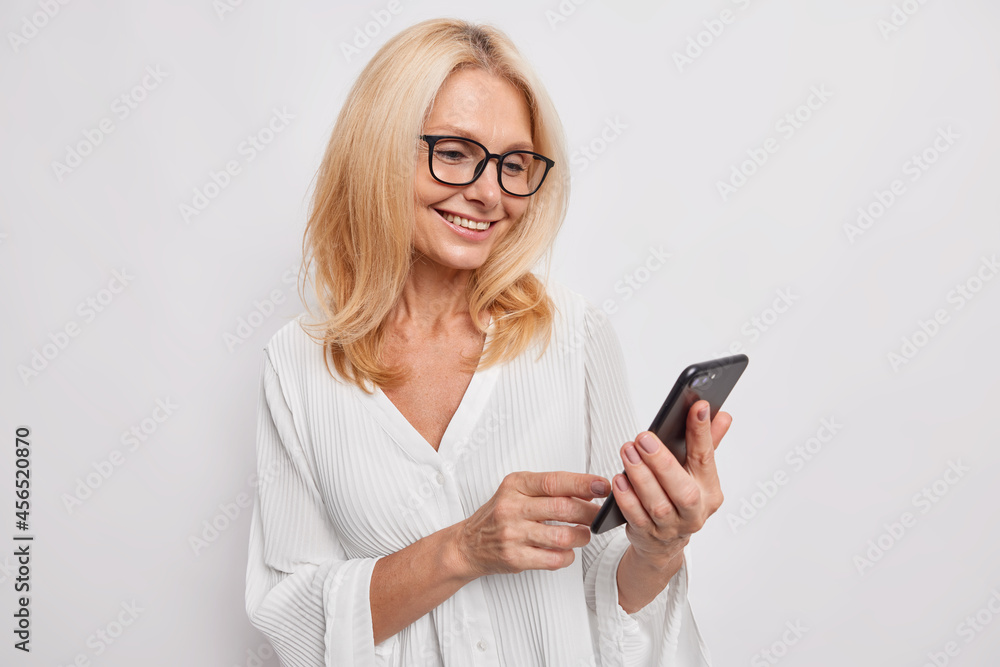 Beautiful middle aged woman chats on mobile phone glad to receive message from daughter smiles happily wears neat white blouse and eyeglasses. Mature businesswoman browses wireless internet. Beautiful middle aged woman chats on mobile phone glad to receive message from daughter smiles happily wears neat white blouse and eyeglasses. Mature businesswoman browses wireless internet.