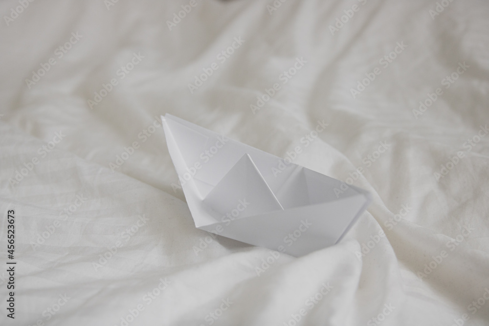 paper origami boat in bed with waves made with bed sheets, natural white crumpled blanket - obrazy, fototapety, plakaty 