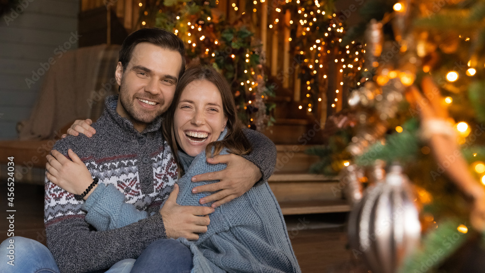 Portrait of happy emotional young affectionate family couple in warm winter sweaters cuddling in decorated living room, feeling excited of celebrating Christmas or New year together, holidays mood.
