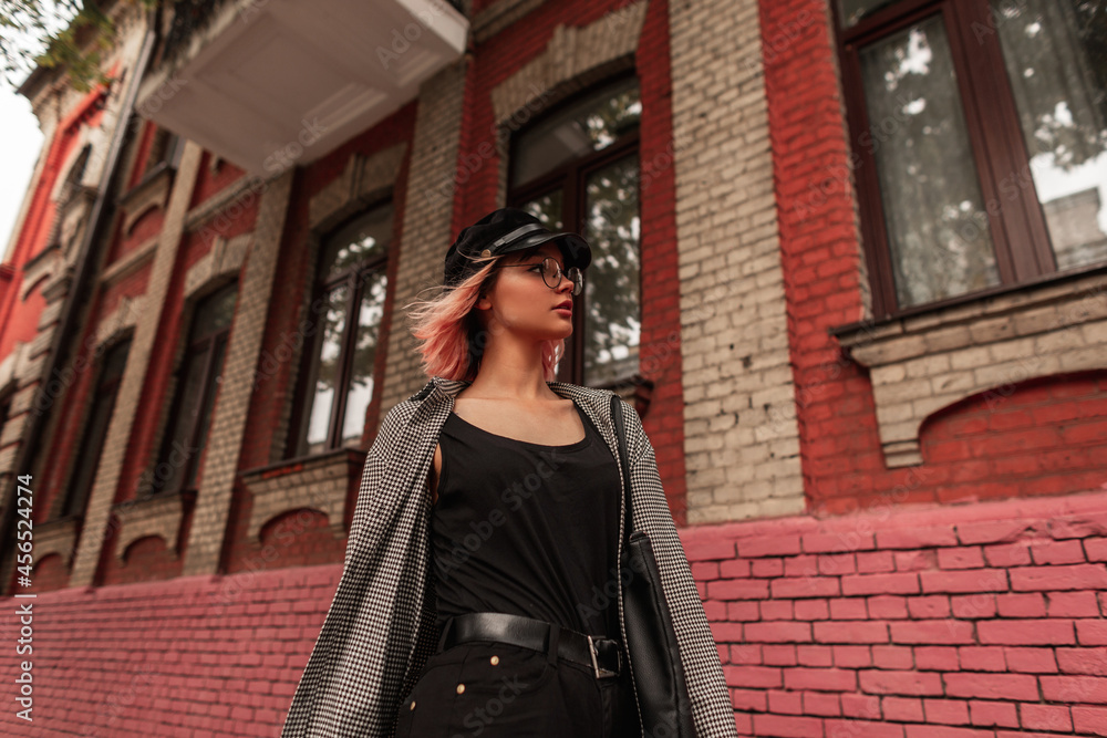 Stylish pretty young model woman with fashionable glasses and a hat in casual outfit with a black t-shirt, plaid shirt and jeans walks on the street near a red brick building