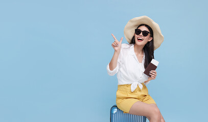 Happy smiling Asian woman dressed in summer clothes and wearing hat with luggage enjoying their summer vacation and pointing finger a copy space on bright blue background..