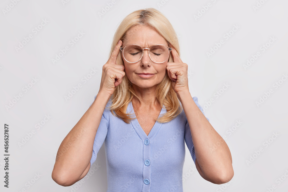 Senior woman has headache touches temples with closed eyes and tensed face suffers from painful migraine wears big optical glasses and blue jumper isolated over white background tries to focus Senior woman has headache touches temples with closed eyes and tensed face suffers from painful migraine wears big optical glasses and blue jumper isolated over white background tries to focus