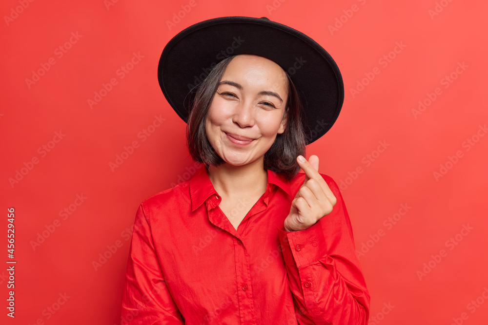 Pretty young Asian woman makes korean like sign smiles gently expresses love wears black hat and shirt poses against vivid red background. Be my valentine. People ethnicity body language concept Pretty young Asian woman makes korean like sign smiles gently expresses love wears black hat and shirt poses against vivid red background. Be my valentine. People ethnicity body language concept