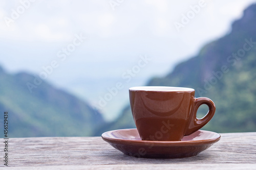 A brown coffee cup on a wooden table with a beautiful scenic view of mountains. Morning coffee feeling happy and free. Space for text