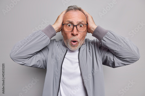 Stupefied impressed bearded man keeps hands on head stares shocked at camera reacts on terrible news dressed in jacket isolated over grey background. Omg concept. Starled scared elderly male model photo