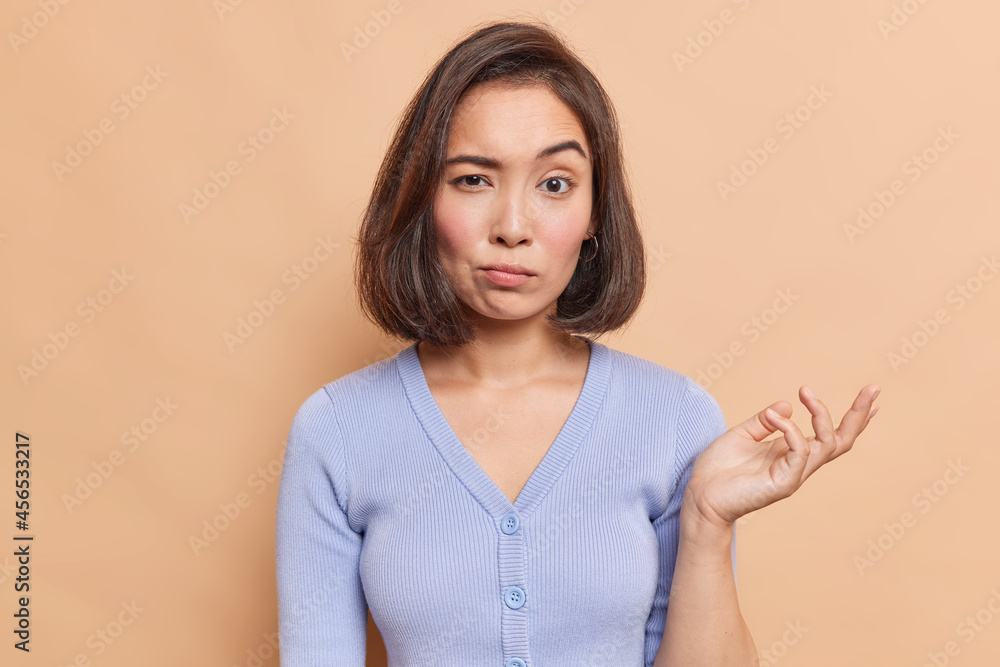 Clueless young Asian woman raises palm with hesitation cannot decide what to do looks unsure dressed in casual blue jumper isolated over beige background has some doubts has confused expression Clueless young Asian woman raises palm with hesitation cannot decide what to do looks unsure dressed in casual blue jumper isolated over beige background has some doubts has confused expression