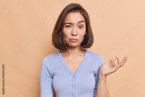 Clueless young Asian woman raises palm with hesitation cannot decide what to do looks unsure dressed in casual blue jumper isolated over beige background has some doubts has confused expression