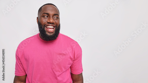Handsome bearded man with dark skin dressed in casual pink t shirt laughs carefree shows optimism poses against grey studio wall blank copy space for your advertisement. Happy emotions concept