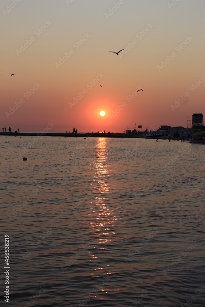 Sunset paints the sky and sea in shades of red. Seagulls fly against the backdrop of the setting sky and the setting sun. The sea pier is visible in the distance. The resting people watch the sunset
