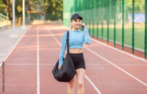 Young athletic woman with training bag going to workout and talking on the phone at the stadium