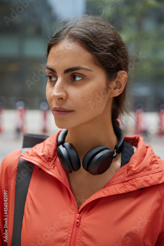 Close up shot of young pensive woman dressed in sportclothes walks with headphones around neck going to practice yoga asana outside thinks how to maintain physical health returns from intense session photo