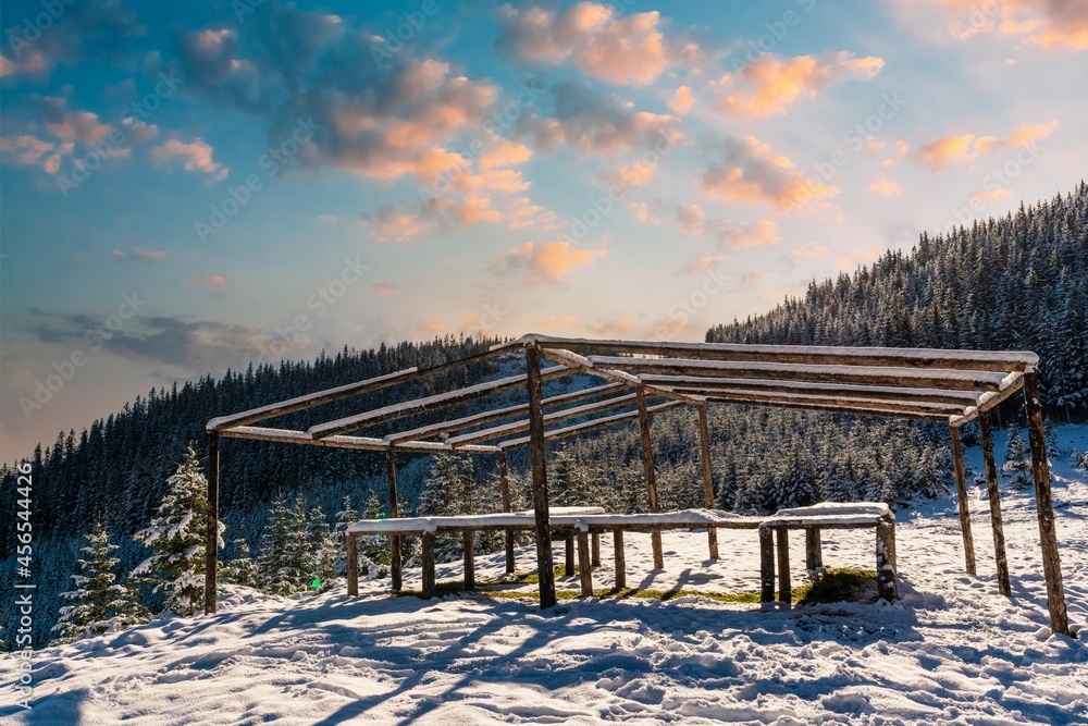 The gazebo on the top of the mountain stands in a snow-covered meadow, bathed in the light of the bright cold sun