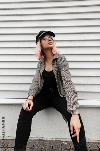 Fashion beautiful young model woman with vintage glasses in trendy casual clothes sitting near a wooden wall