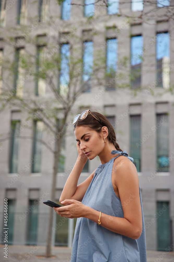 Vertical shot of beautiful woman holds mobile phone reads notification about transaction surfs internet wears sunglasses casual t shirt strolls in city with modern technologies messages on summer day