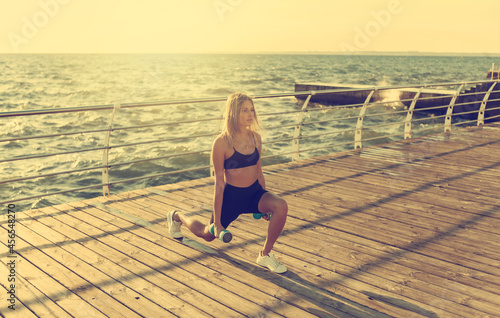 Young slim woman in sportswear practicing lunges with dumbbells in hands on the beach at sunrise. Morning workout.