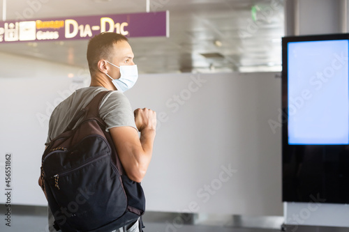 Young European man in gray coat, protective disposable medical mask in airport. Afraid of dangerous N-CoV 2019 influenza coronavirus. backpack, suitcase.