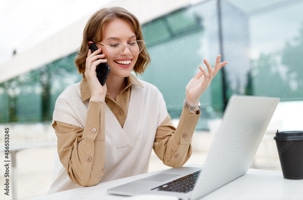Office employee talking on cell phone with smile sitting on terrace in cafe with coffee and laptop Office employee talking on cell phone with smile sitting on terrace in cafe with coffee and laptop