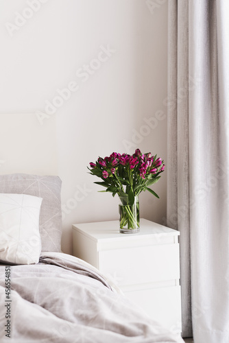 Simple home decor: fresh Alstroemeria flowers in a glass vase on a bedside table of white color in a bedroom. Cozy and comfortable atmosphere. Minimal style in interior design