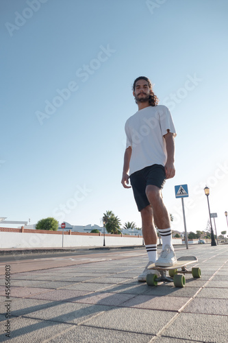 Young attractive stylish caucasian italian skater standing on the street. He is looking confident at camera and wearing summer clothes.