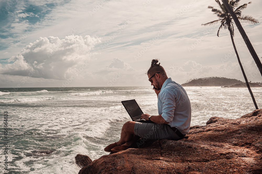 Cyfrowy nomada, człowiek pracujący z laptopem i smartfonem zdalnie na tle oceanu i skał siedzący na wybrzeżu. - obrazy, fototapety, plakaty 