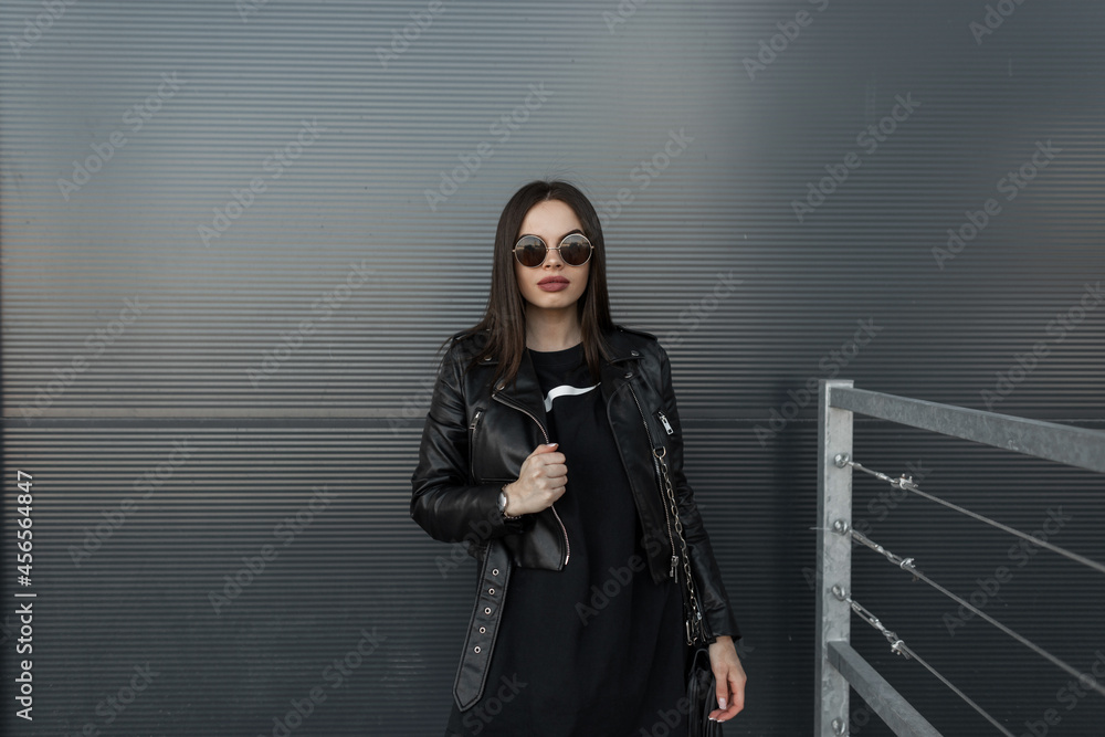 Stylish pretty hipster woman in fashionable round sunglasses and leather black jacket with sweatshirt stands near metal wall in city Stylish pretty hipster woman in fashionable round sunglasses and leather black jacket with sweatshirt stands near metal wall in city
