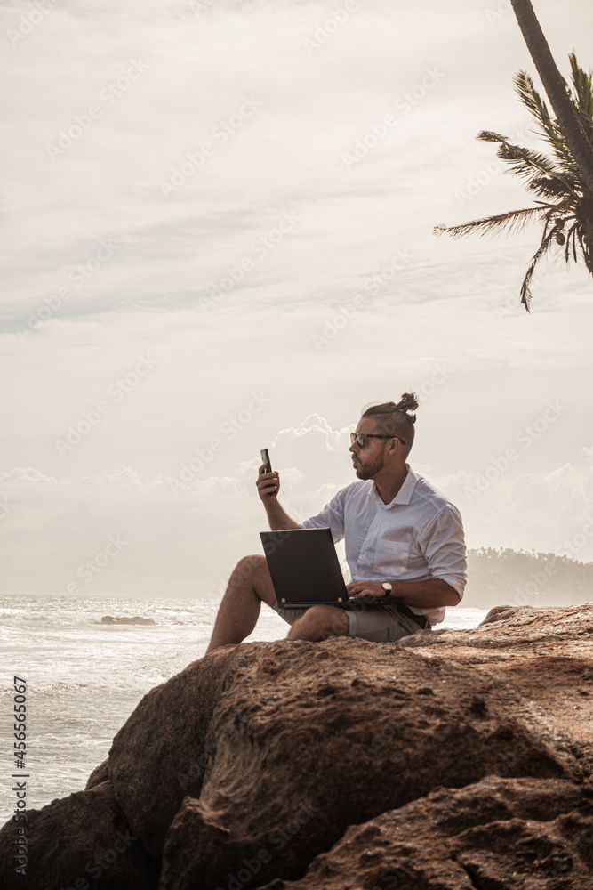 Cyfrowy nomada, człowiek pracujący z laptopem i smartfonem zdalnie na tle oceanu i skał siedzący na wybrzeżu. - obrazy, fototapety, plakaty 