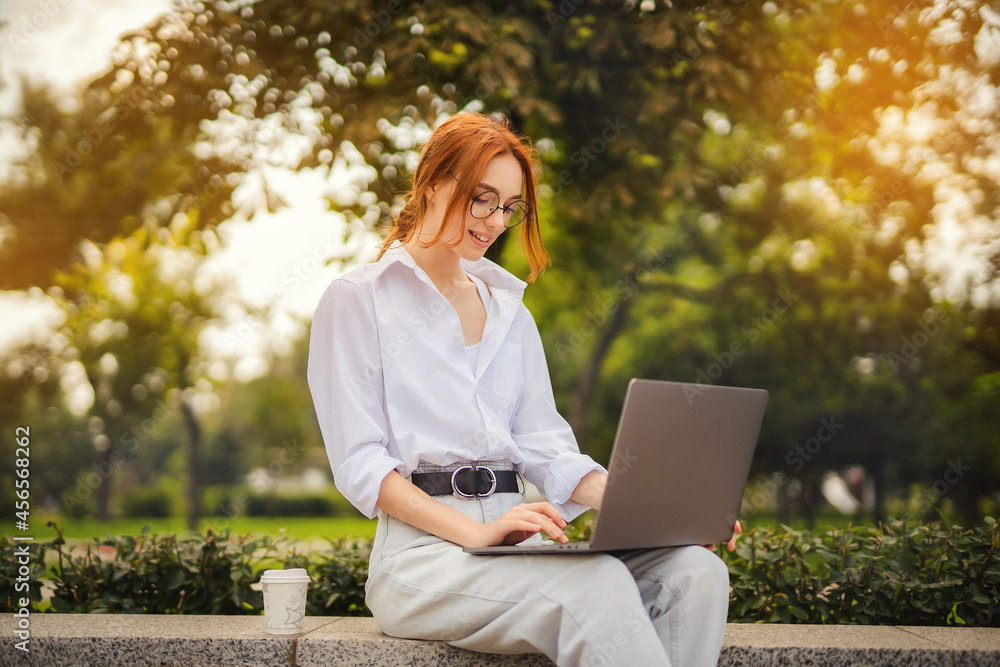 Beautiful redhead young woman sitting in the park and using laptop. Student. University. Freelance. Wearing in glasses casual jeans and white shirt. Coffee to go. Paper cup. Looking in laptop Beautiful redhead young woman sitting in the park and using laptop. Student. University. Freelance. Wearing in glasses casual jeans and white shirt. Coffee to go. Paper cup. Looking in laptop