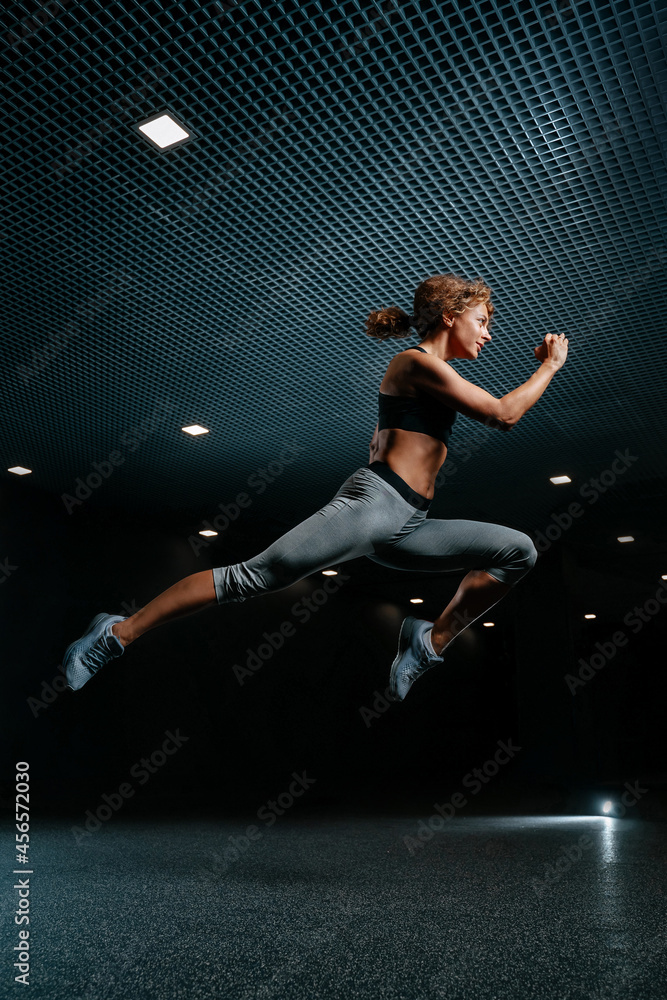 Slender woman is actively jumping high in sports clothes on a black background of the gym. Dynamic movement side view.
