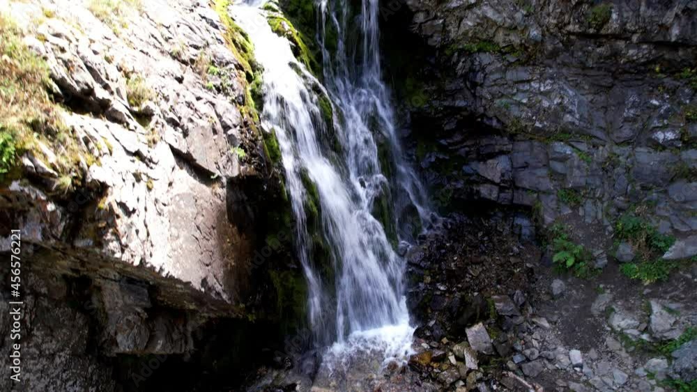 A mountain waterfall in a rocky gorge. The view from the drone is of steep cliffs, moss, green plants and the river. There are stones lying. Splashes from the water. There is a log lying. Almaty A mountain waterfall in a rocky gorge. The view from the drone is of steep cliffs, moss, green plants and the river. There are stones lying. Splashes from the water. There is a log lying. Almaty