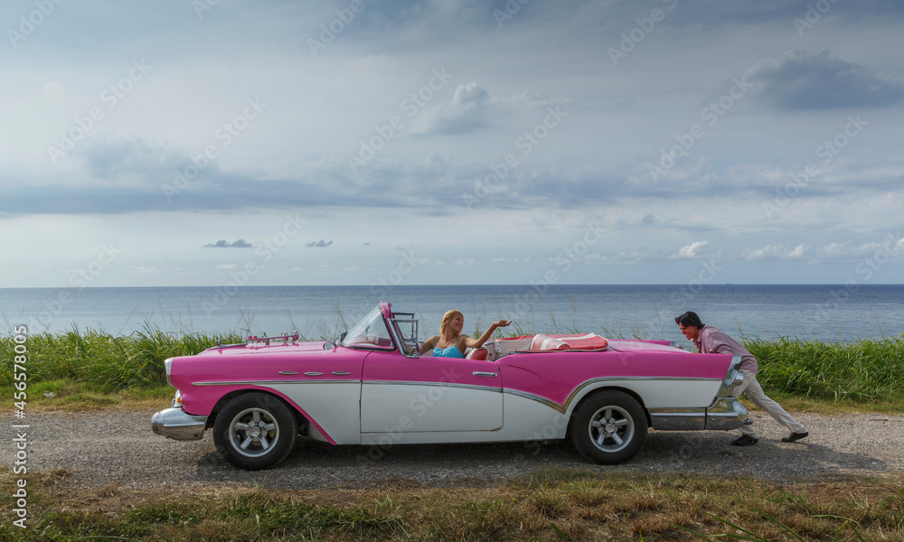 Young man pushing vintage convertible at coast whilst girlfriend drives, Havana, Cuba - obrazy, fototapety, plakaty 