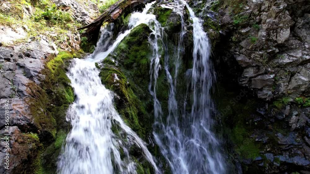 A mountain waterfall in a rocky gorge. The view from the drone is of steep cliffs, moss, green plants and the river. There are stones lying. Splashes from the water. There is a log lying. Almaty A mountain waterfall in a rocky gorge. The view from the drone is of steep cliffs, moss, green plants and the river. There are stones lying. Splashes from the water. There is a log lying. Almaty