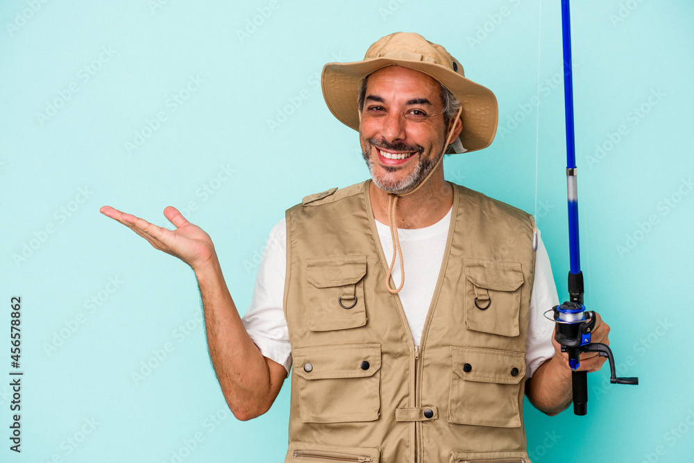 Middle age caucasian fisherman holding rod isolated on blue background showing a copy space on a palm and holding another hand on waist. Middle age caucasian fisherman holding rod isolated on blue background showing a copy space on a palm and holding another hand on waist.