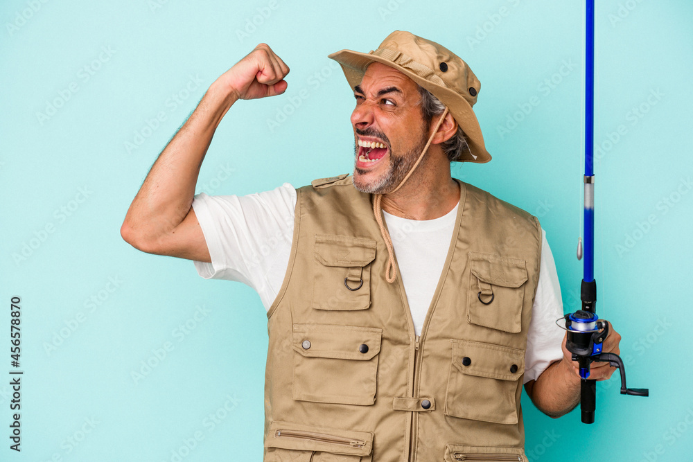 Middle age caucasian fisherman holding rod isolated on blue background  raising fist after a victory, winner concept.
