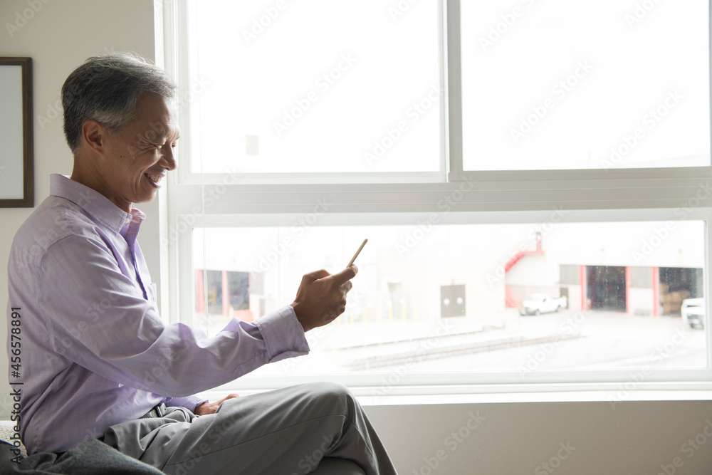 Side view of mature man sitting in front of window using smartphone