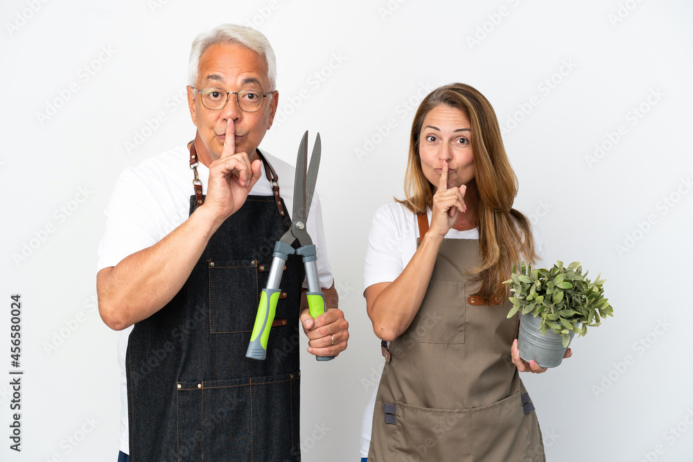 Middle age gardeners holding a plant and scissors isolated on white background showing a sign of silence gesture putting finger in mouth Middle age gardeners holding a plant and scissors isolated on white background showing a sign of silence gesture putting finger in mouth