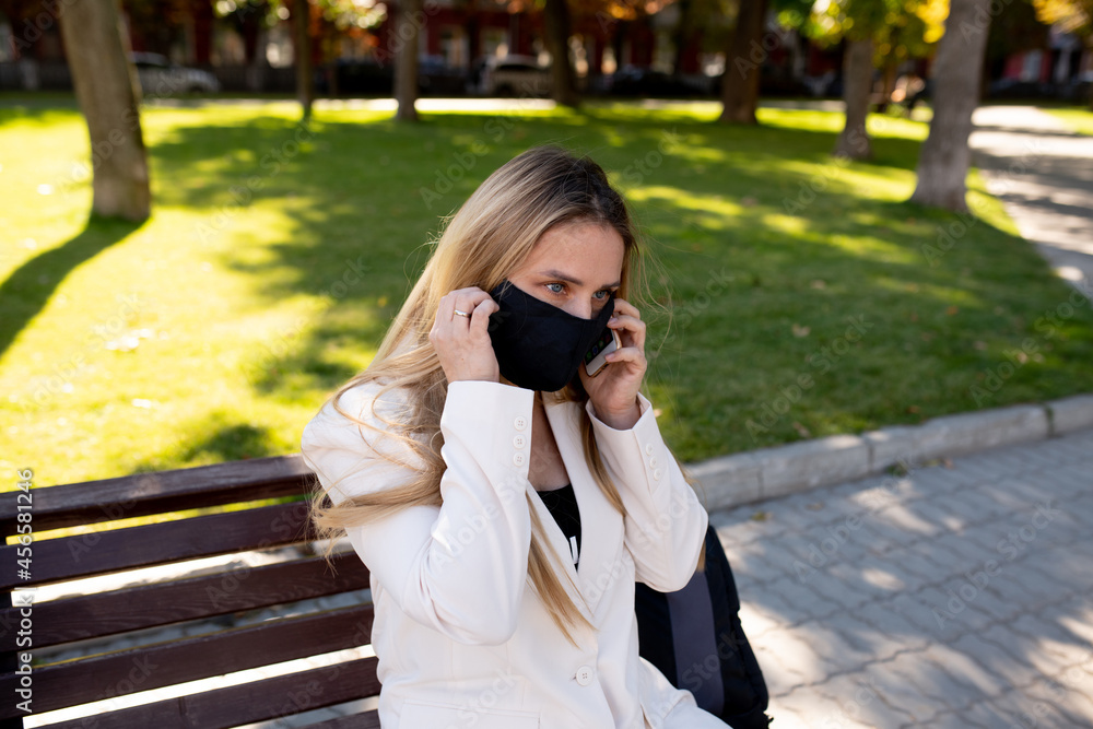 Young woman wearing a medical protective mask sits on a park bench during the coronavirus pandemic and talks on a mobile video
