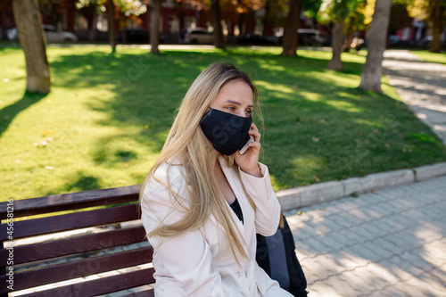Young woman wearing a medical protective mask sits on a park bench during the coronavirus pandemic and talks on a mobile video