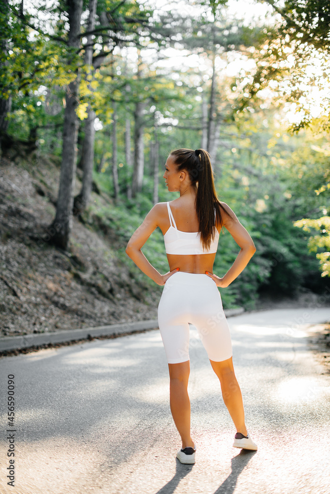 A young beautiful girl poses with her back before running training, on the road in a dense forest, during sunset. A healthy lifestyle and running in the fresh air. A young beautiful girl poses with her back before running training, on the road in a dense forest, during sunset. A healthy lifestyle and running in the fresh air.