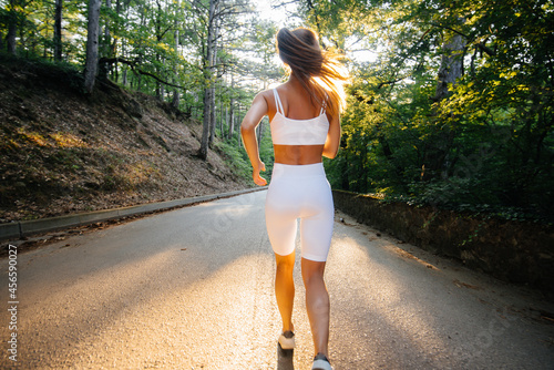A young beautiful girl in white sports clothes is running with her back, on the road in a dense forest, during sunset. Doing sports in the fresh air. A healthy lifestyle.