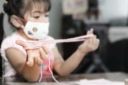 Little Asian girl wearing facial hygienic mask playing cats cradle game at home selective focus. Quarantine Home isolation during COVID-19 pandemic.