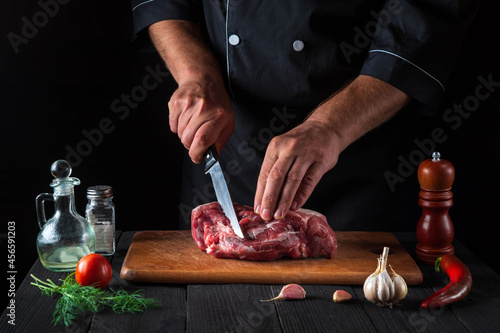 Professional chef cuts meat with a knife in the kitchen prepares food. Vegetables and spices on the kitchen table in a restaurant to prepare a delicious lunch