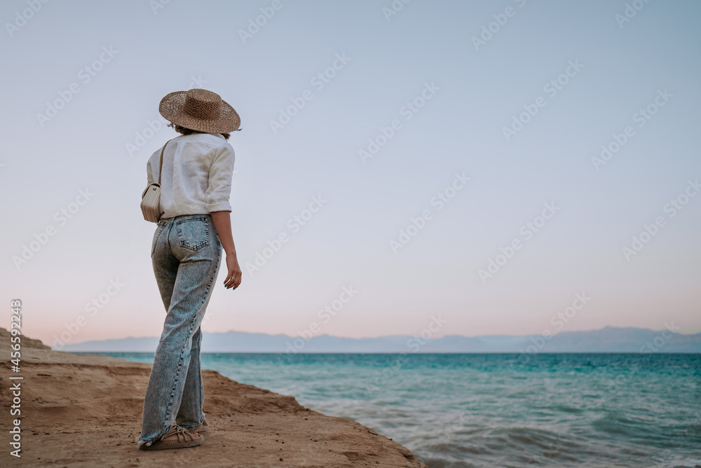 Stranger woman standing on beach near Mediterranean Ionian sea. Lady tourist in straw hat watching beautiful blue water surface alone, nature background. Windy weather, golden hour.