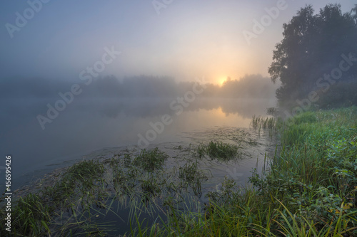 Sunrise over the foggy lake with the reflection of sun. Mist on the water, forest silhouettes and the rays of the rising sun. Beautiful morning landscape with sunrise over river.