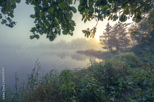 Sunrise over the foggy lake with the reflection of sun. Mist on the water  forest silhouettes and the rays of the rising sun. Beautiful morning landscape with sunrise over river.