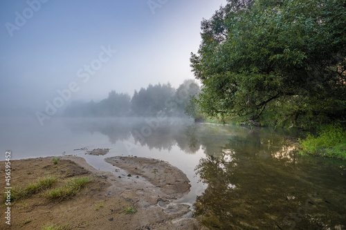Sunrise over the foggy lake with the reflection of sun. Mist on the water, forest silhouettes and the rays of the rising sun. Beautiful morning landscape with sunrise over river.