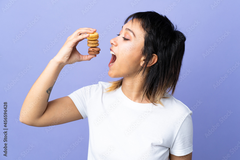 Young Uruguayan woman over isolated purple background holding colorful French macarons and eating it Young Uruguayan woman over isolated purple background holding colorful French macarons and eating it