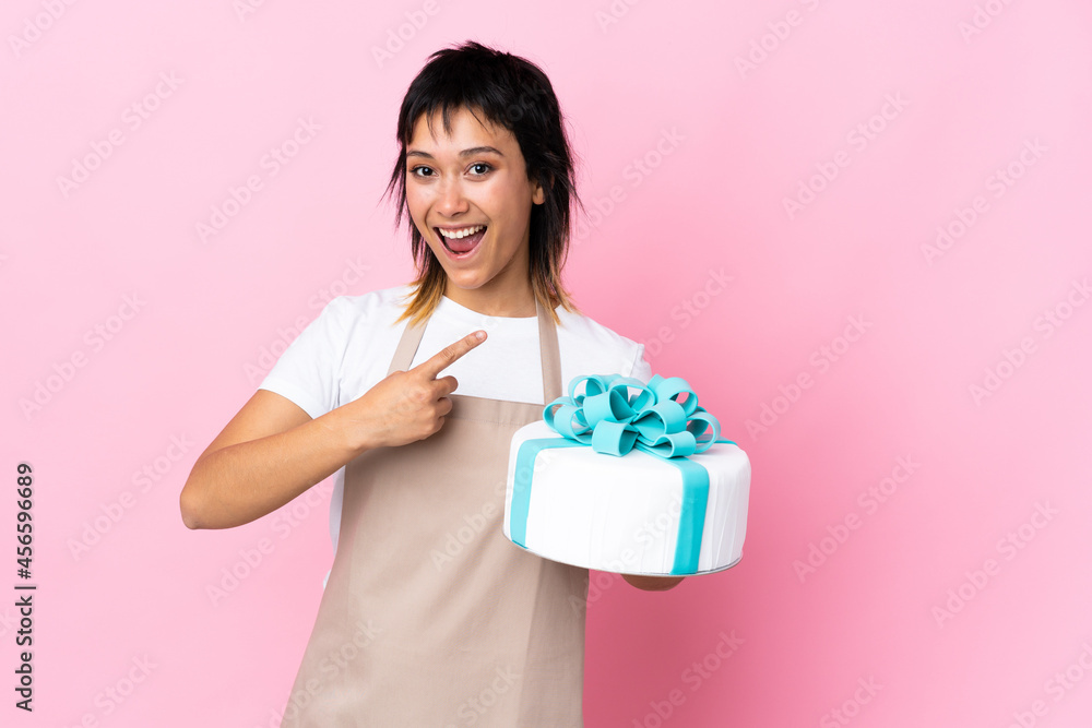Uruguayan Pastry chef holding a big cake over isolated pink background pointing finger to the side Uruguayan Pastry chef holding a big cake over isolated pink background pointing finger to the side