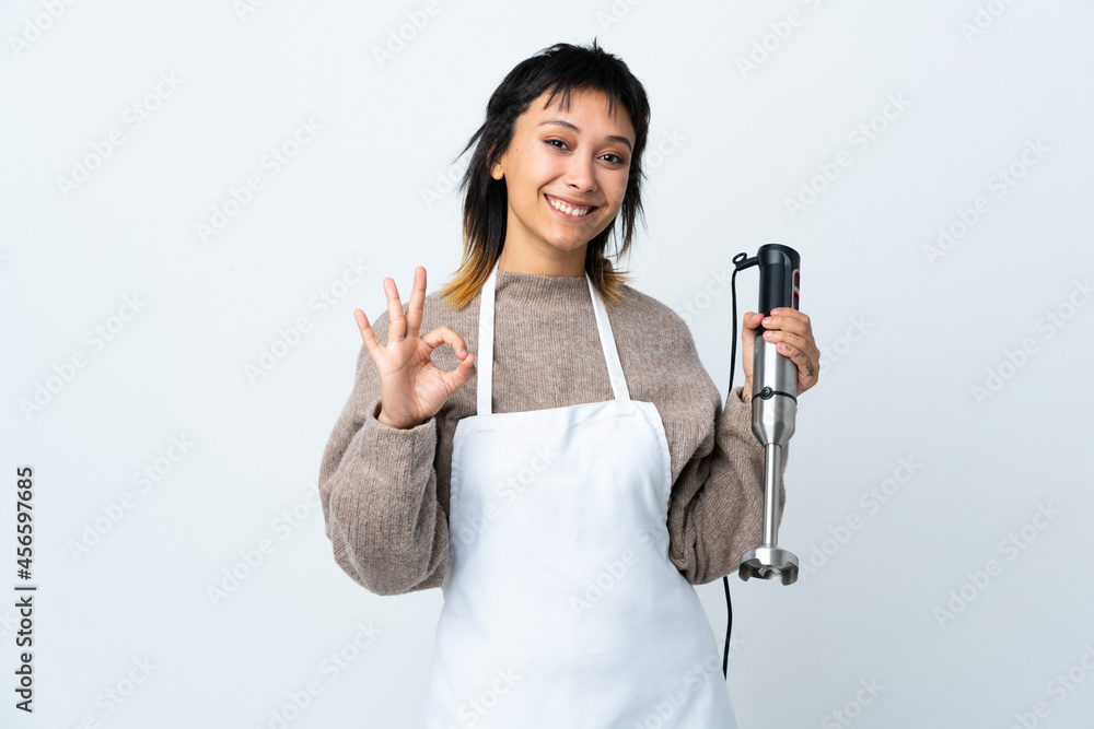 Chef Uruguayan girl using hand blender over isolated white background showing an ok sign with fingers Chef Uruguayan girl using hand blender over isolated white background showing an ok sign with fingers