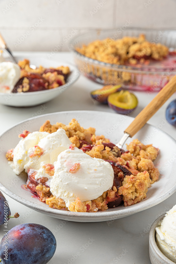 Fruit crumble. Plum crumble pie with vanilla ice cream in plates with spoons on white marble table for tasty breakfast. Fruit crumble. Plum crumble pie with vanilla ice cream in plates with spoons on white marble table for tasty breakfast.