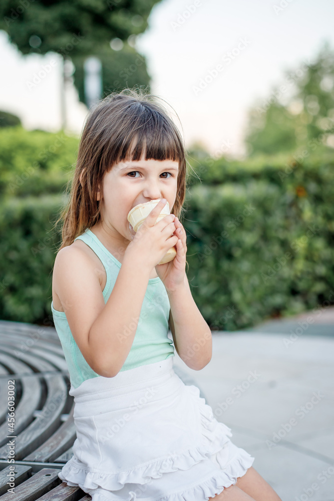 Fototapeta premium girl in a light dress eats popsicle ice cream sitting on a bench in a city park. Dessert.